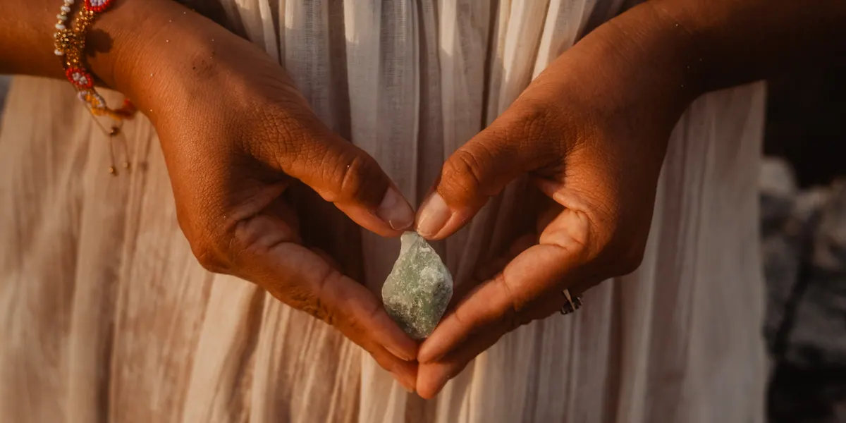Dos manos forman un corazón alrededor de un cuarzo verde, sobre un vestido de lino claro